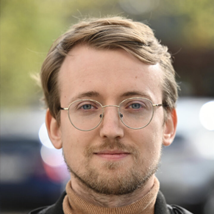 A young, smiling man with blond hair and a beard. He wears glasses.