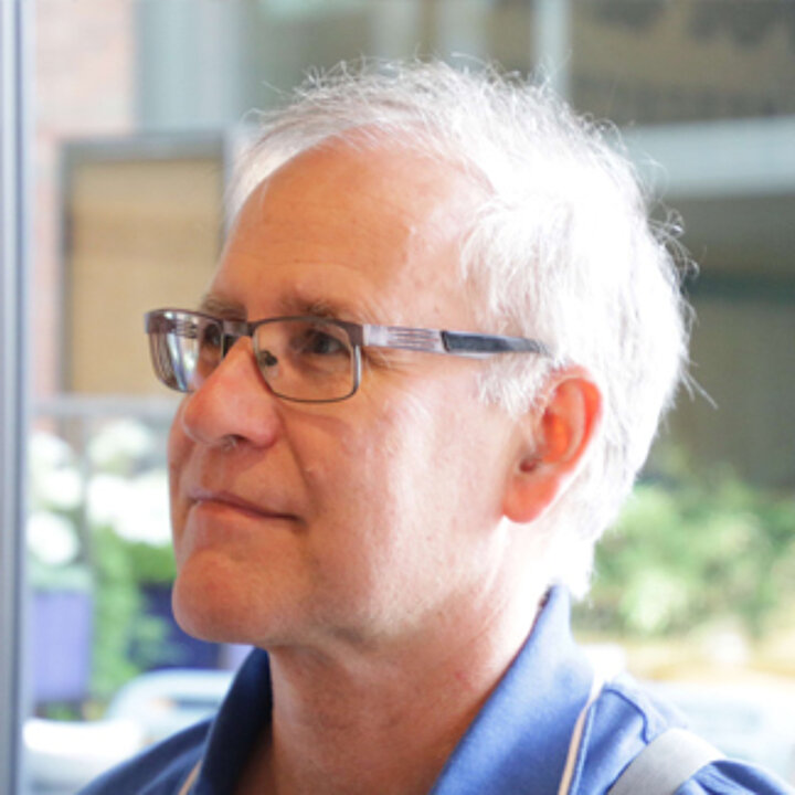 An older, graying man with glasses and a blue shirt. He looks into the distance.